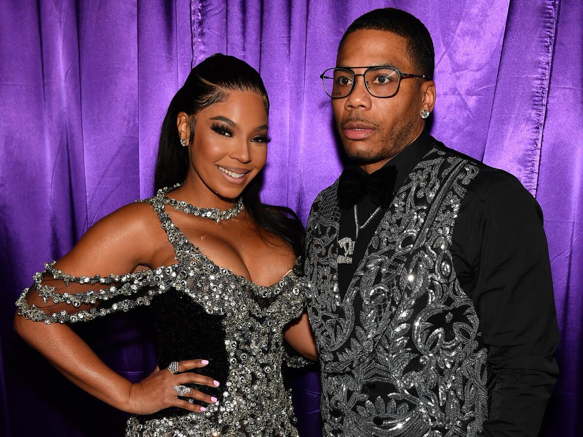 Ashanti smiling in a glittering gown and standing with Nelly, who has on a matching glittery vest, black shirt and glasses