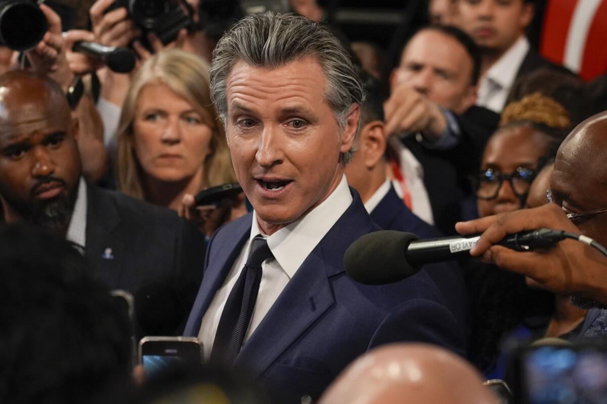 California Gov. Gavin Newsom, center, wearing a dark suit and tie while surrounded by a crush of reporters holding equipment