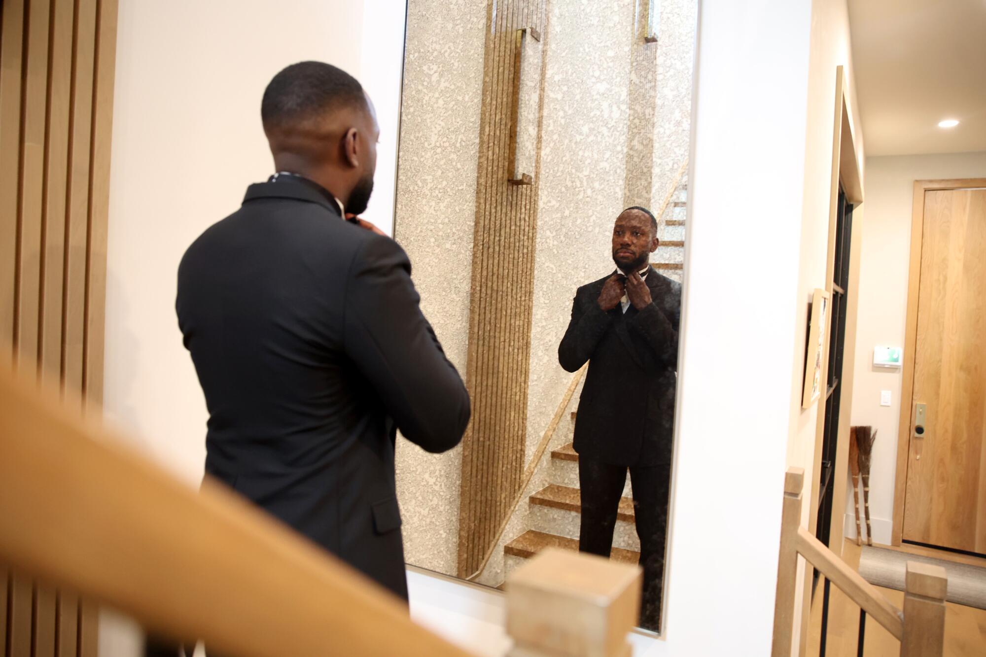 A man in a tuxedo looks at his reflection in a mirror near a stairwell.