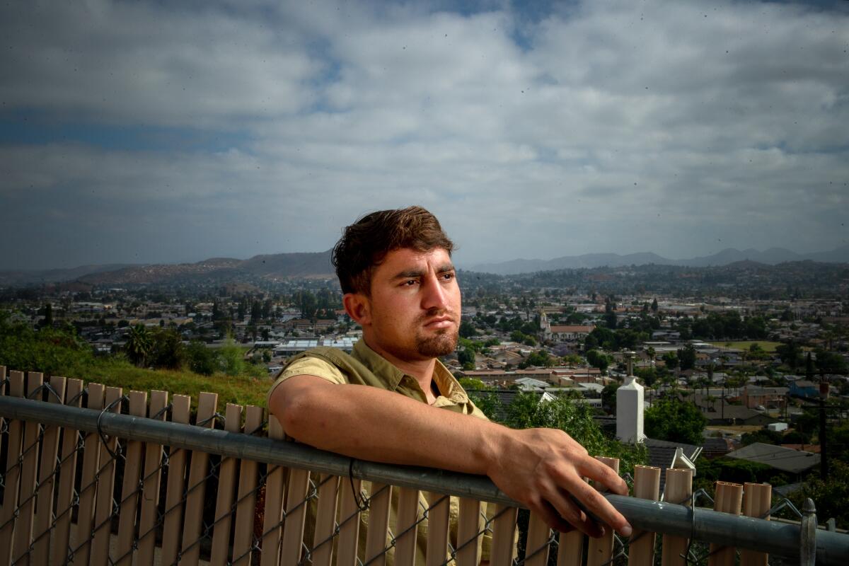 A bearded man stands behind a chain-link fence on a hill with a city in the background.