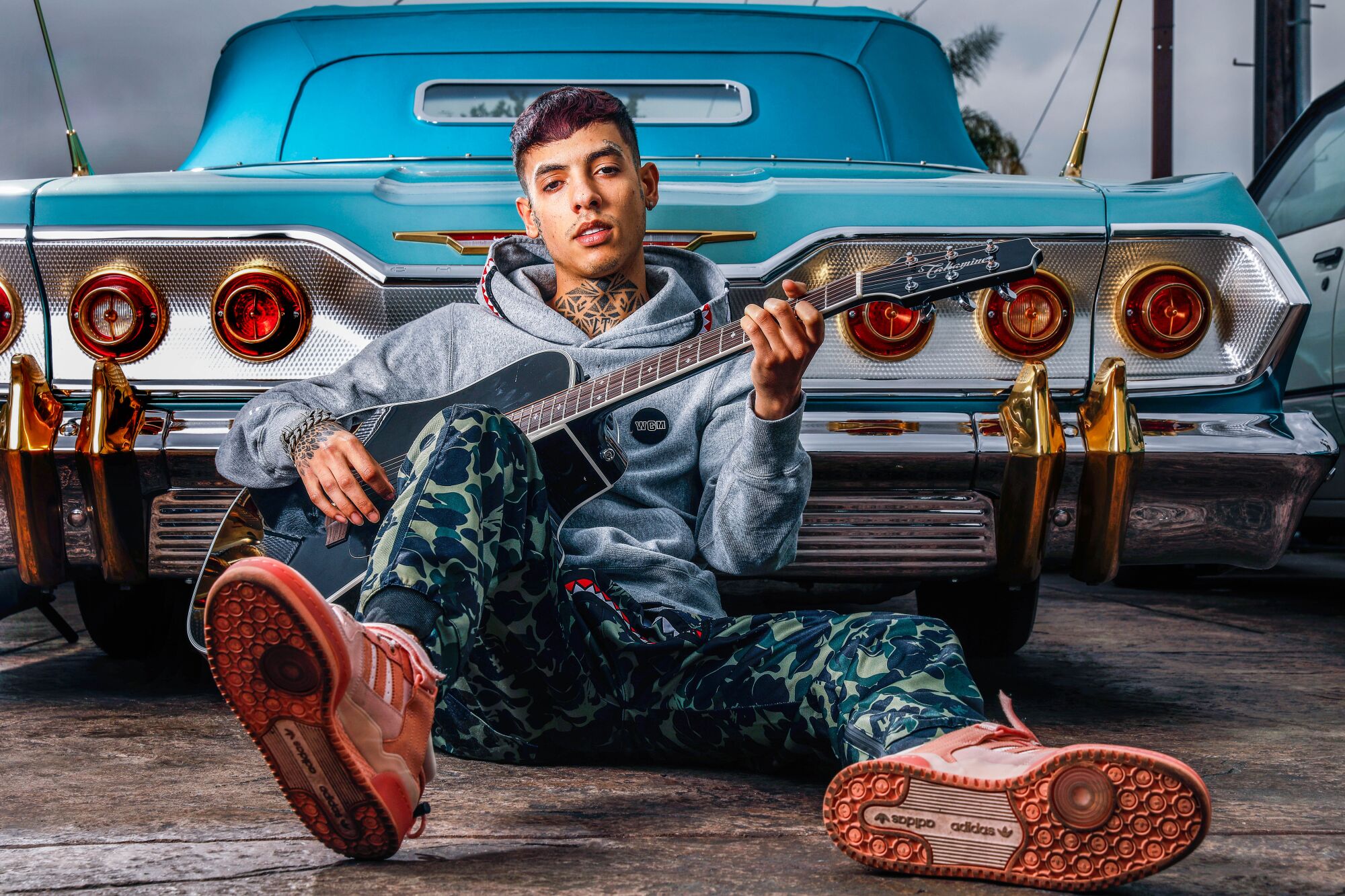 Natanael Cano holds a guitar and leans against a vintage car. 