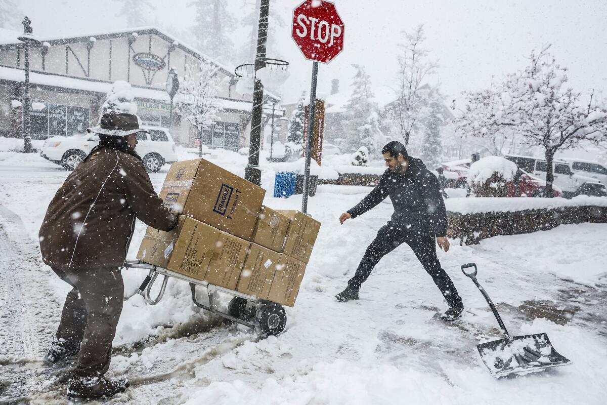 A man shovels a sidewalk of snow as another wheels a dolly loaded with boxes.