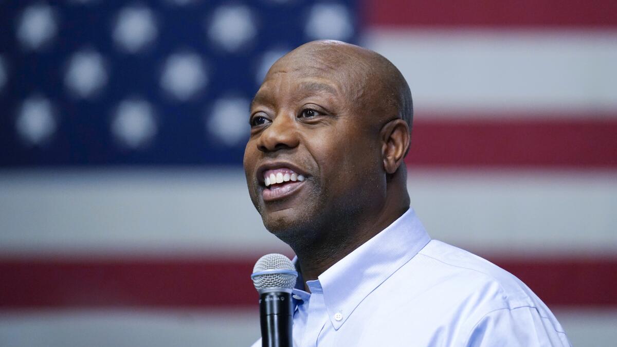 Sen. Tim Scott pictured from the shoulders up, speaking into a microphone in front of a large American flag background