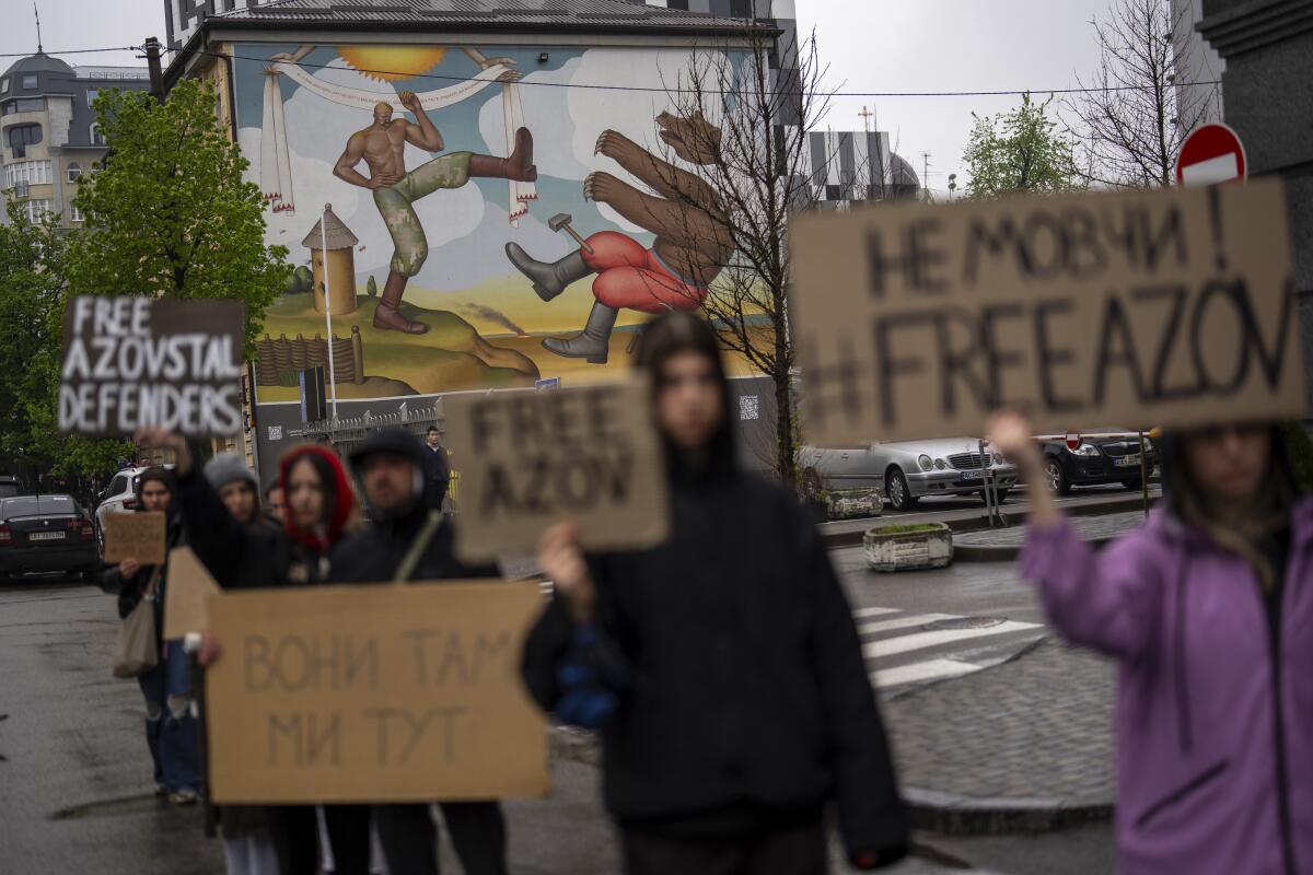 People stand near a Kyiv, Ukraine, mural of a man kicking a bear, holding signs urging release of prisoners of war.