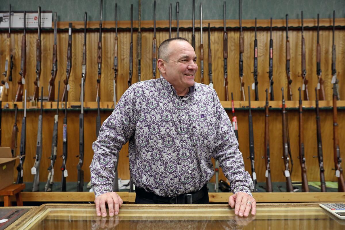 A smiling man with rifles on display behind him.