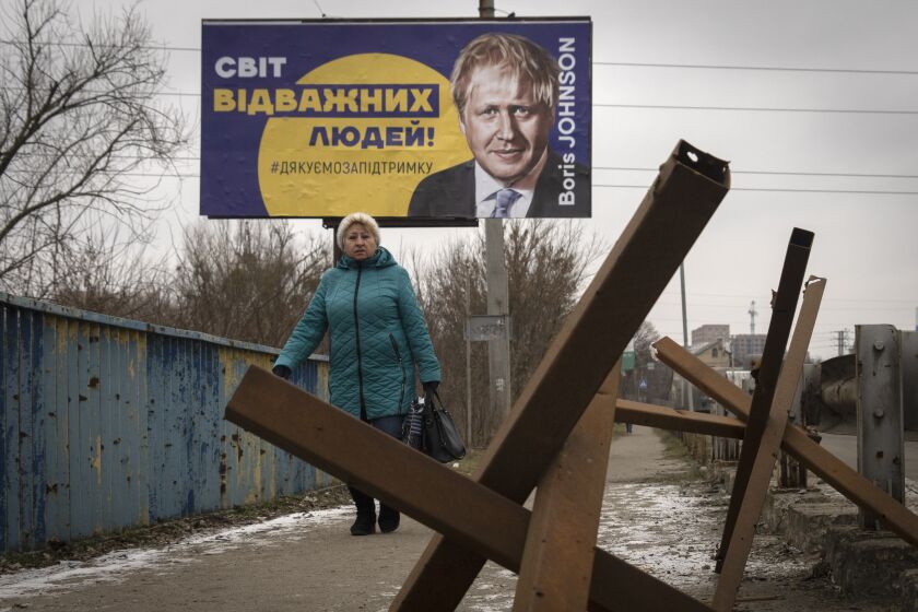 A woman passes by a board depicting former British Prime Minister Boris Johnson and reads: "The World of Brave People! #thank you for support", with antitank hedgehogs in the foreground, in the town of Bucha, outside Kyiv, Ukraine, Monday Jan. 30, 2023. (AP Photo/Efrem Lukatsky)