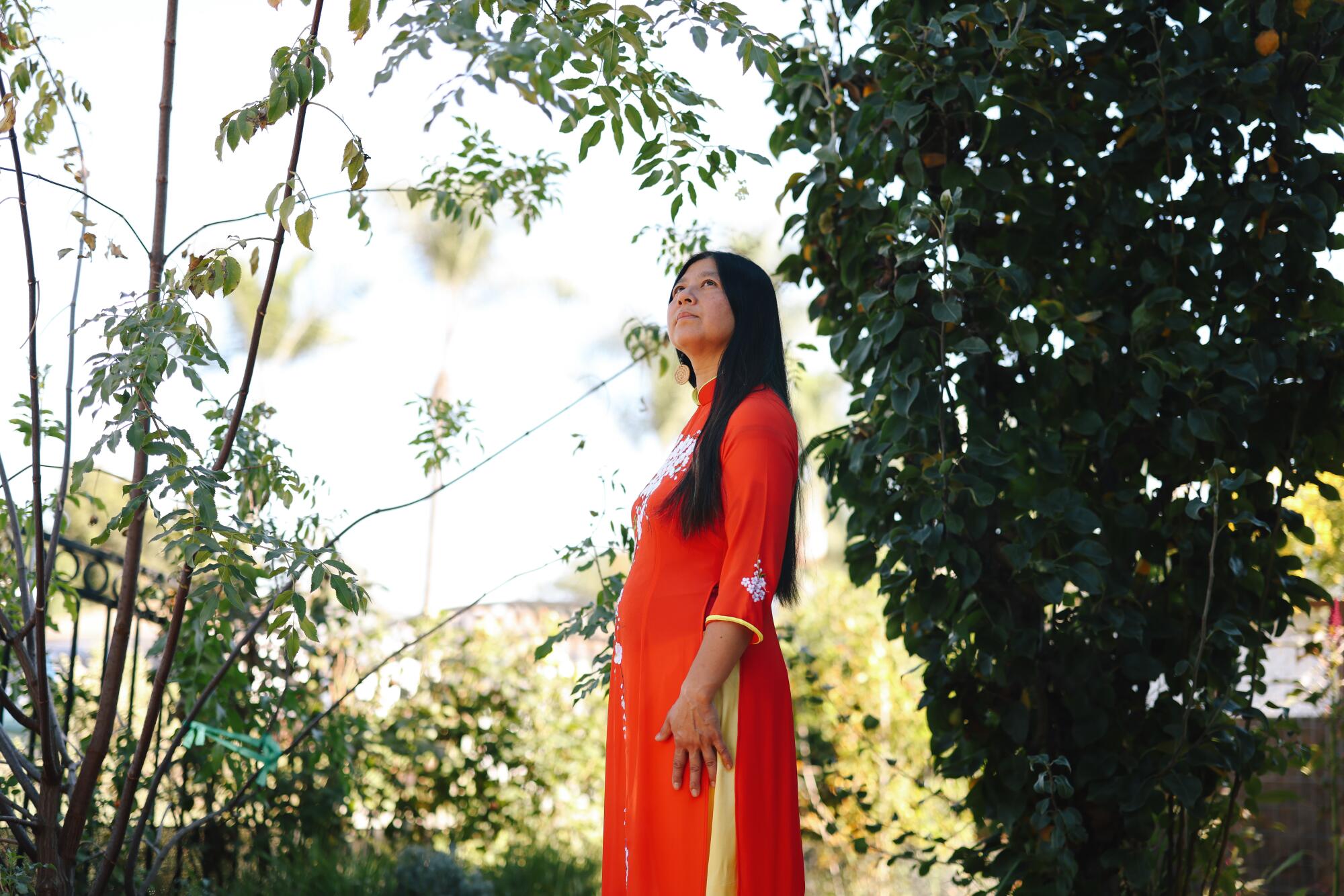Teresa Mei Chuk em um vestido vermelho brilhante olhando para longe entre as árvores verdes. 