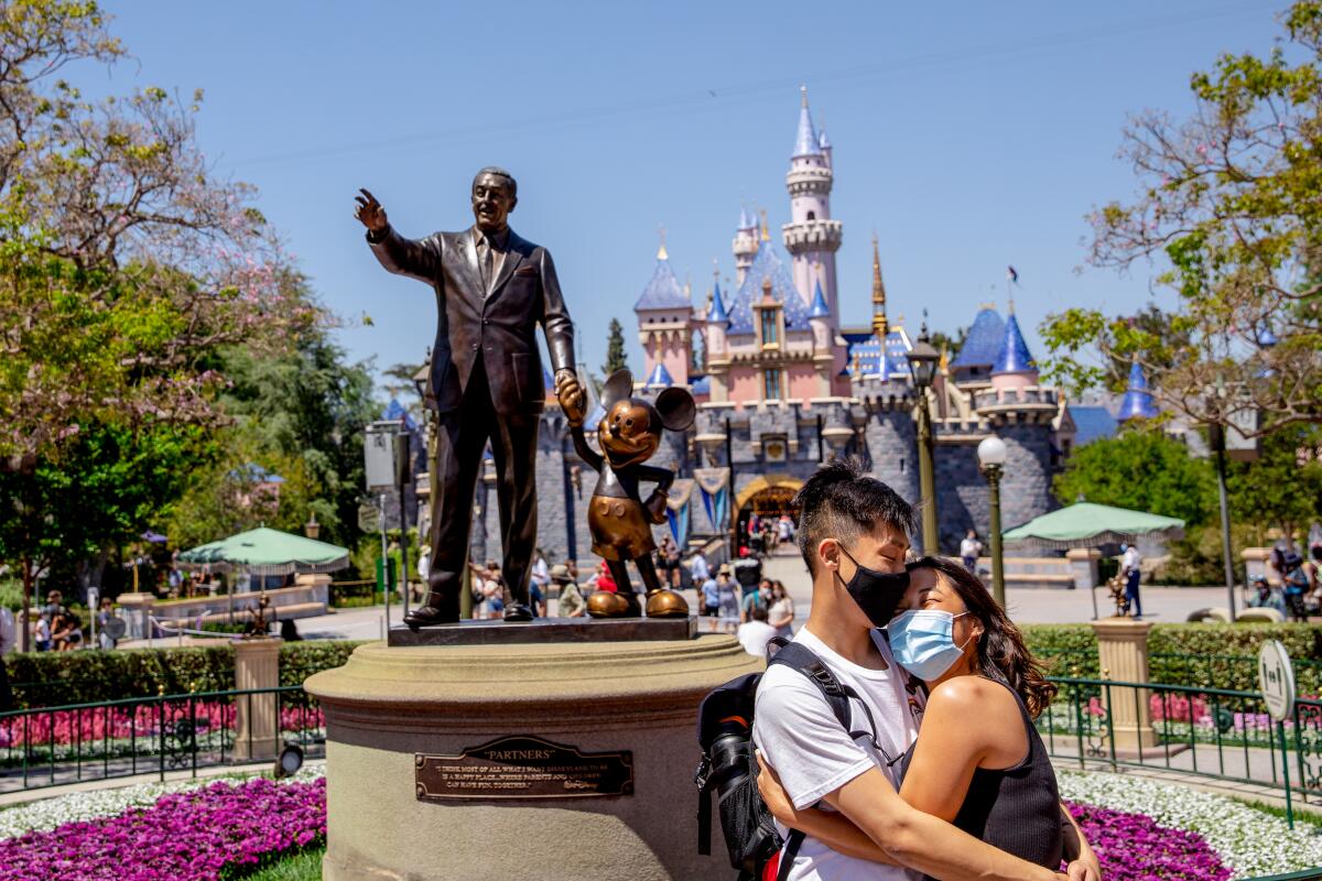 A masked couple hugs in front of the Walt Disney statue at Disneyland.
