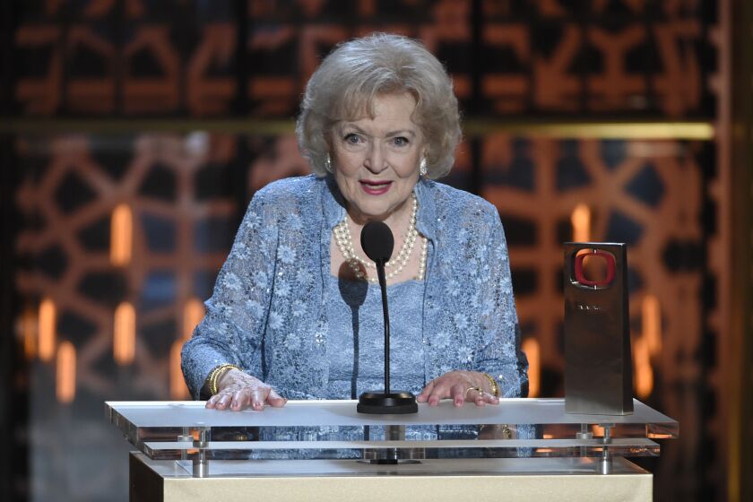 An older woman with white hair in a blue outfit speaks at an awards show lectern