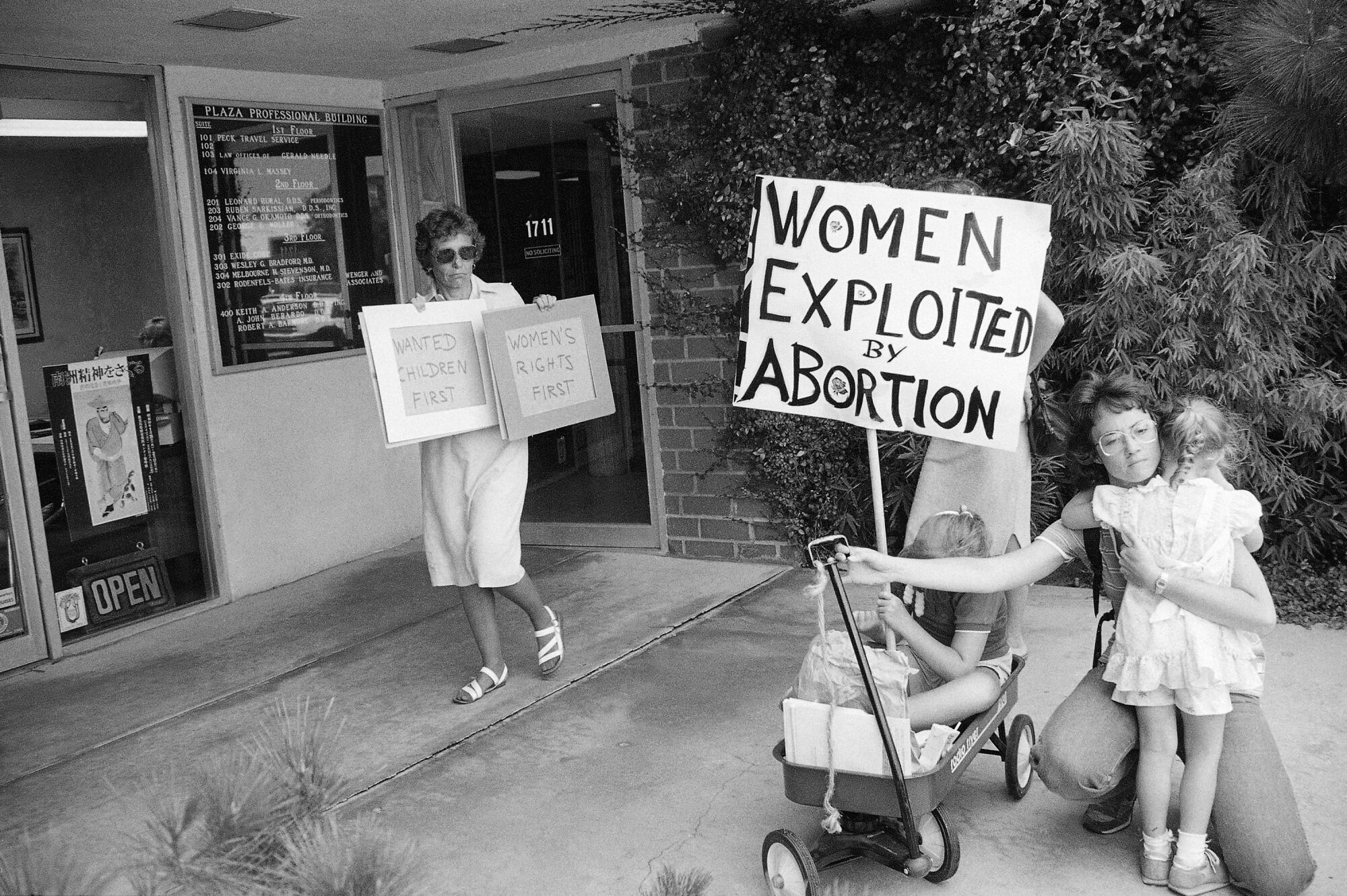 A black-and-white photo of a woman holding two pro-abortion rights signs next to two women and two children protesting