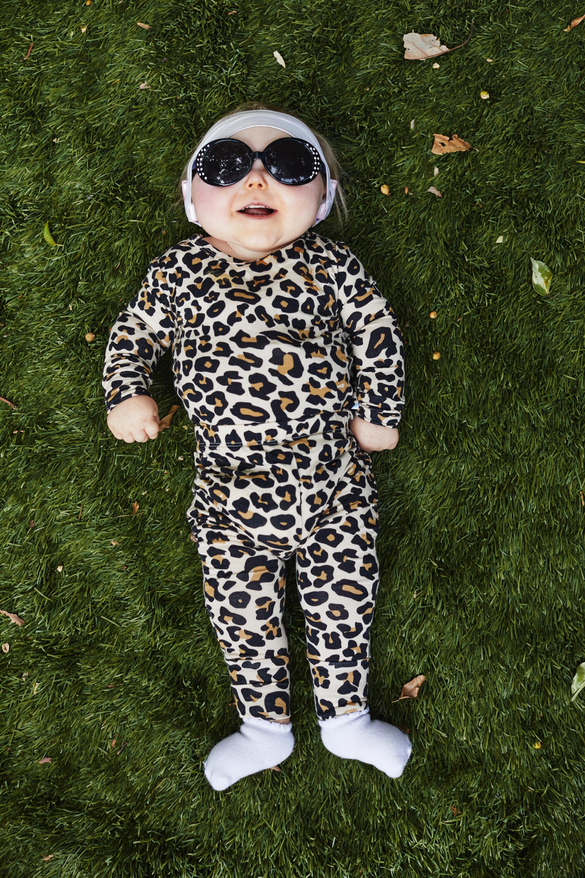 A baby wearing sunglasses and a leopard-print onesie lies on her back on a lawn.