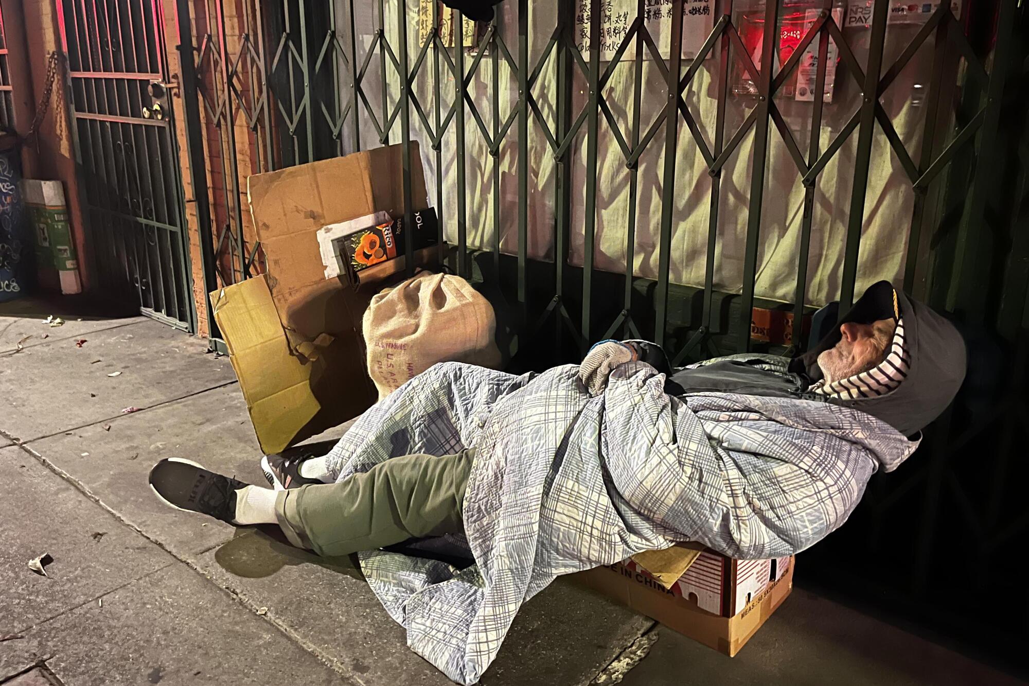 A person sleeps on a cardboard bed on a sidewalk near the gated and locked entry of a business. 