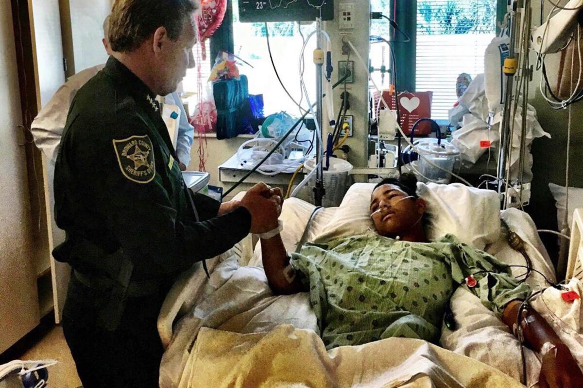 A sheriff  holds the hand of a severely wounded teen in a hospital bed.