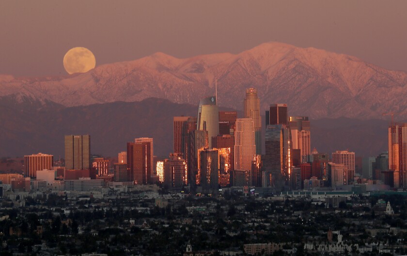 A full moon rises over the snow-capped San Gabriel Mountains and the skyline of downtown Los Angeles.