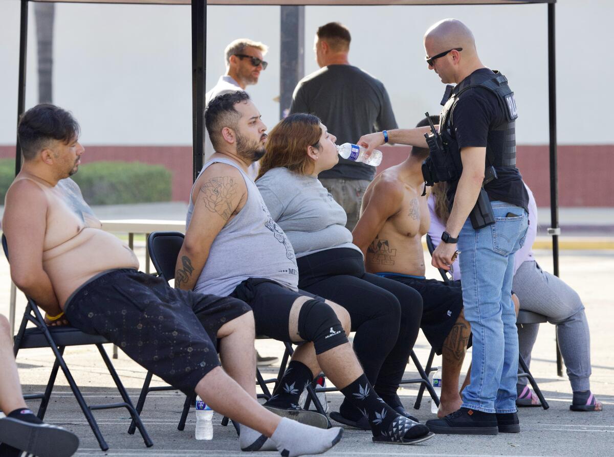 An officer gives a drink of water to one of several seated people in handcuffs.