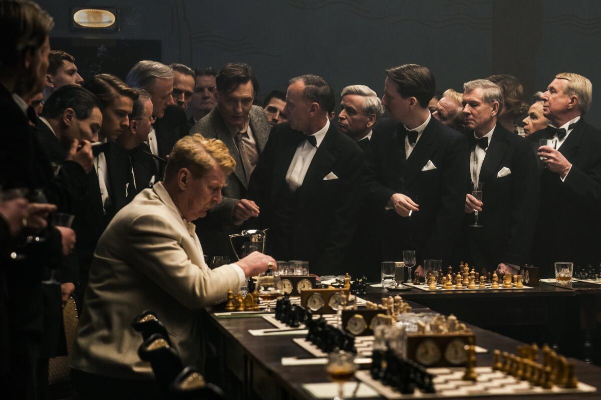 man sits playing chess while a crowd of men in tuxes is near him 
