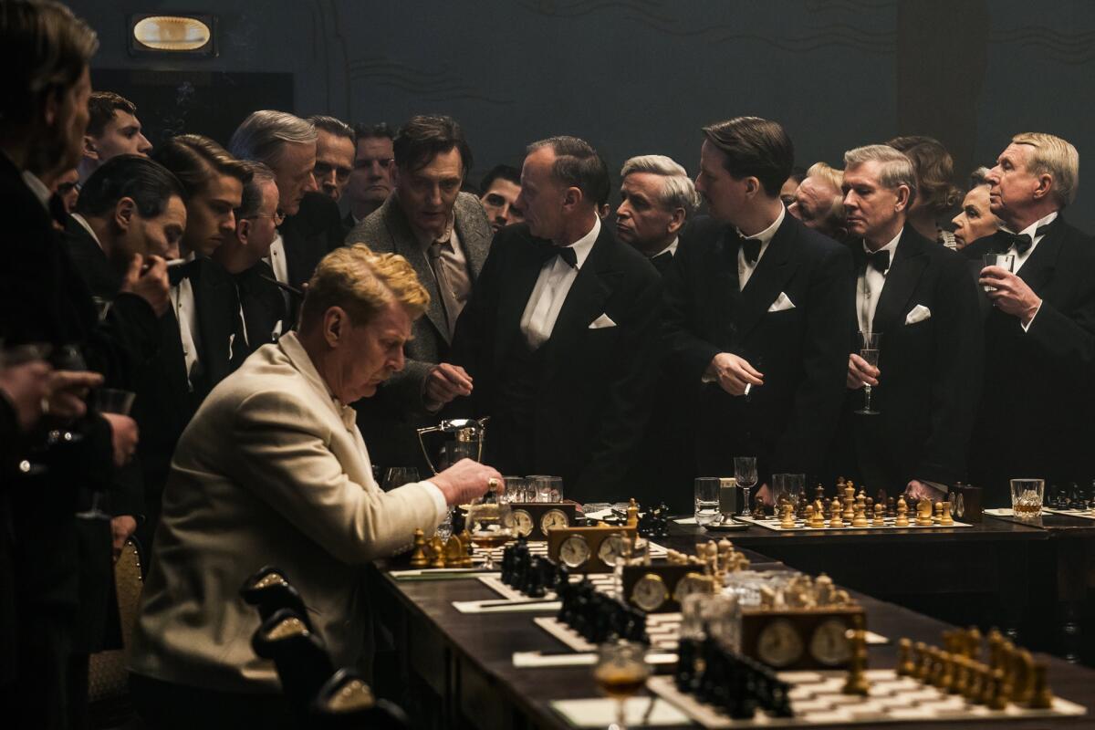 man sits playing chess while a crowd of men in tuxes is near him