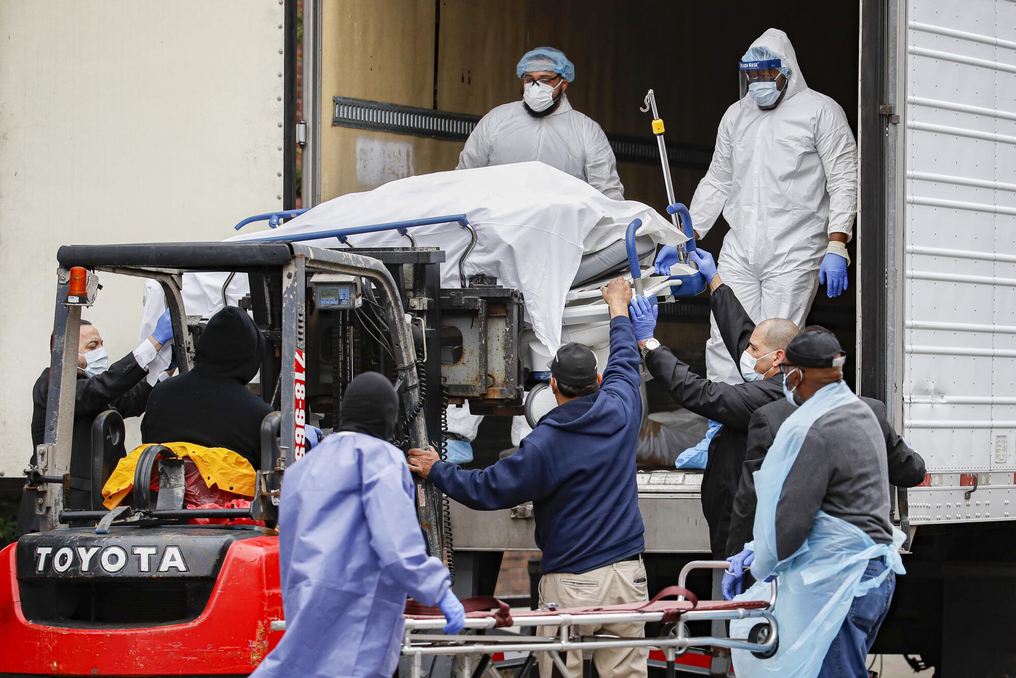 A body wrapped in plastic is unloaded from a refrigerated truck and handled by medical workers 