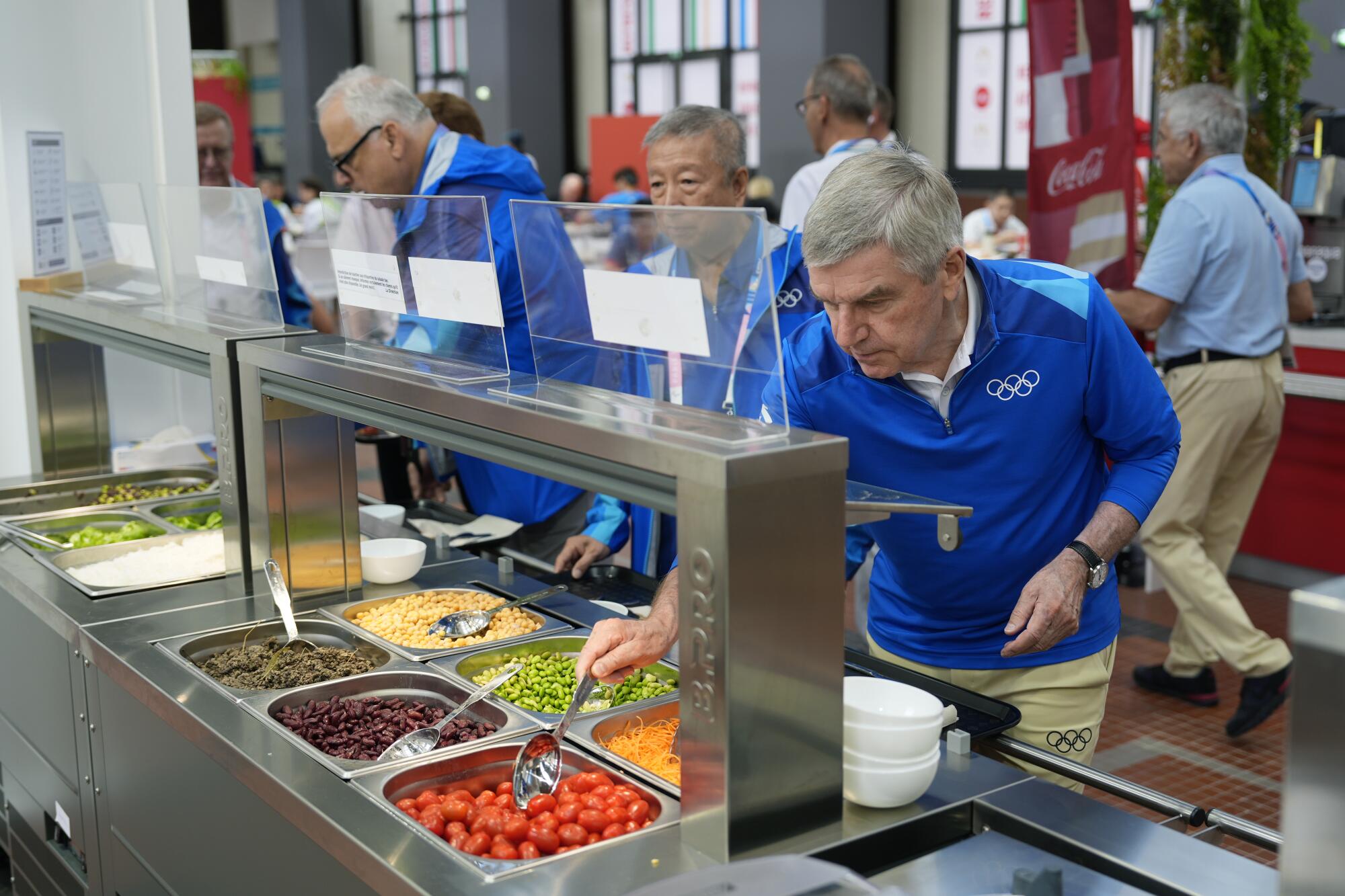 A man in blue uses a spoon to serve himself tomatoes at a salad bar.