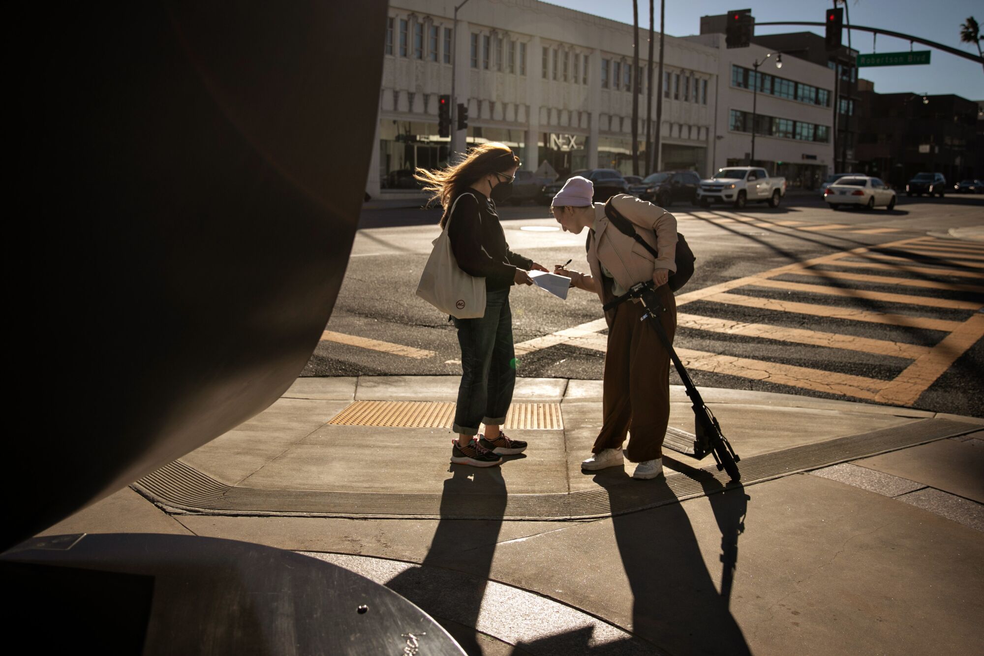 İki kadın kaldırımın köşesinde karşı karşıya dururken, biri diğerinin tuttuğu panodaki bir kağıdı imzalıyor.