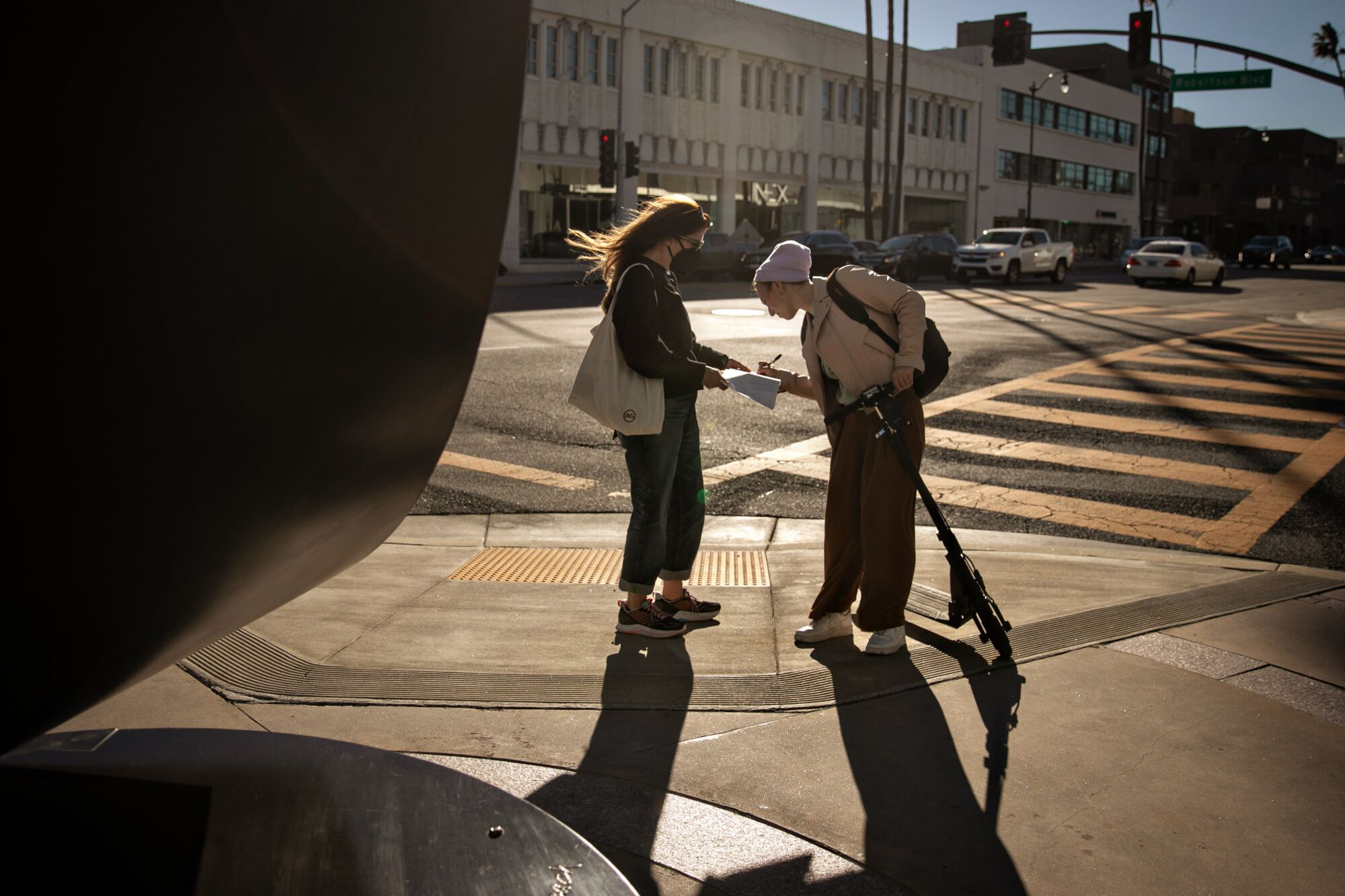 Deux femmes se font face sur un coin de trottoir, tandis que l'une signe un papier sur une planchette tenue par l'autre.