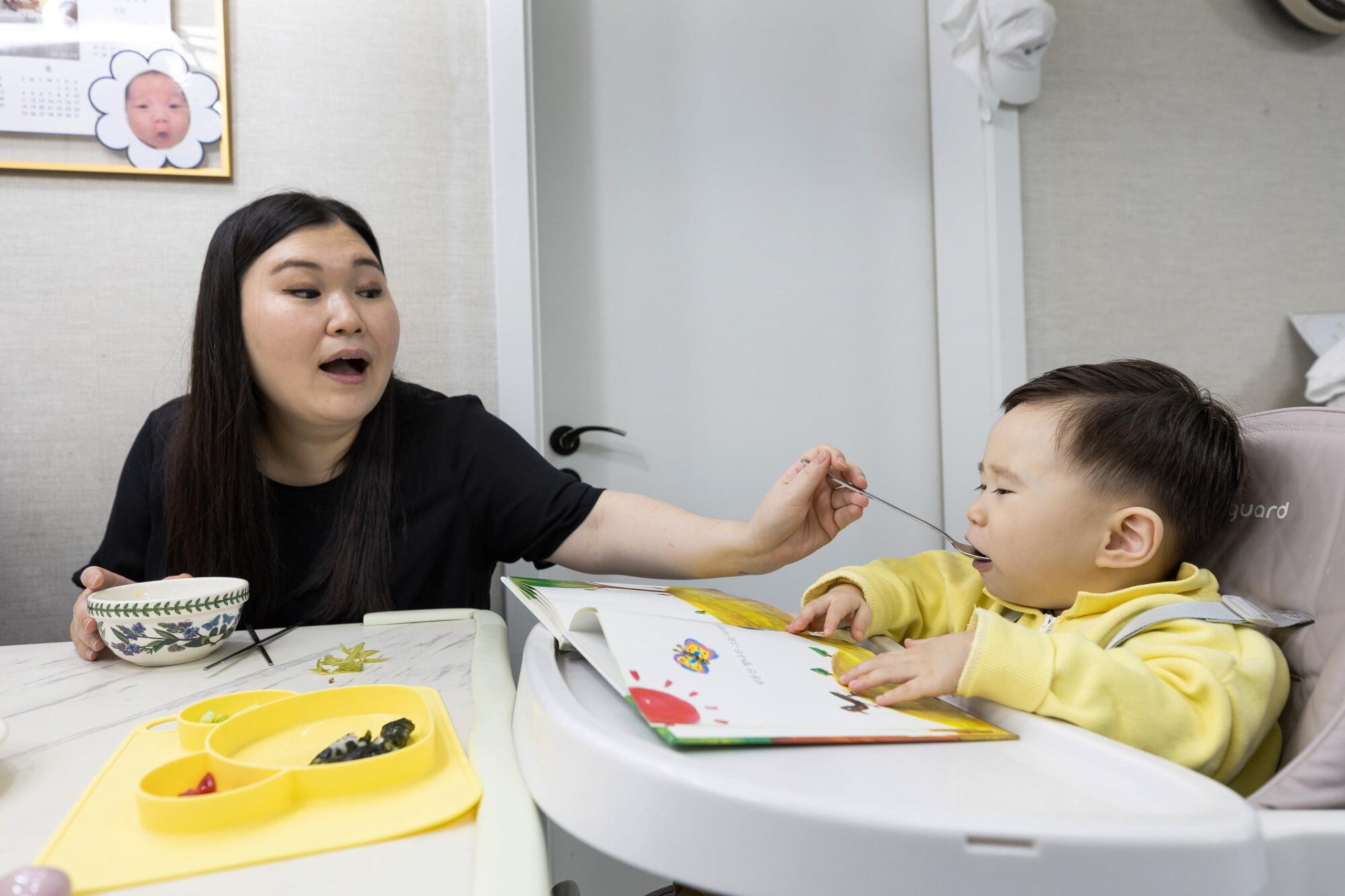 A seated woman puts food in the mouth of a baby in a high chair 