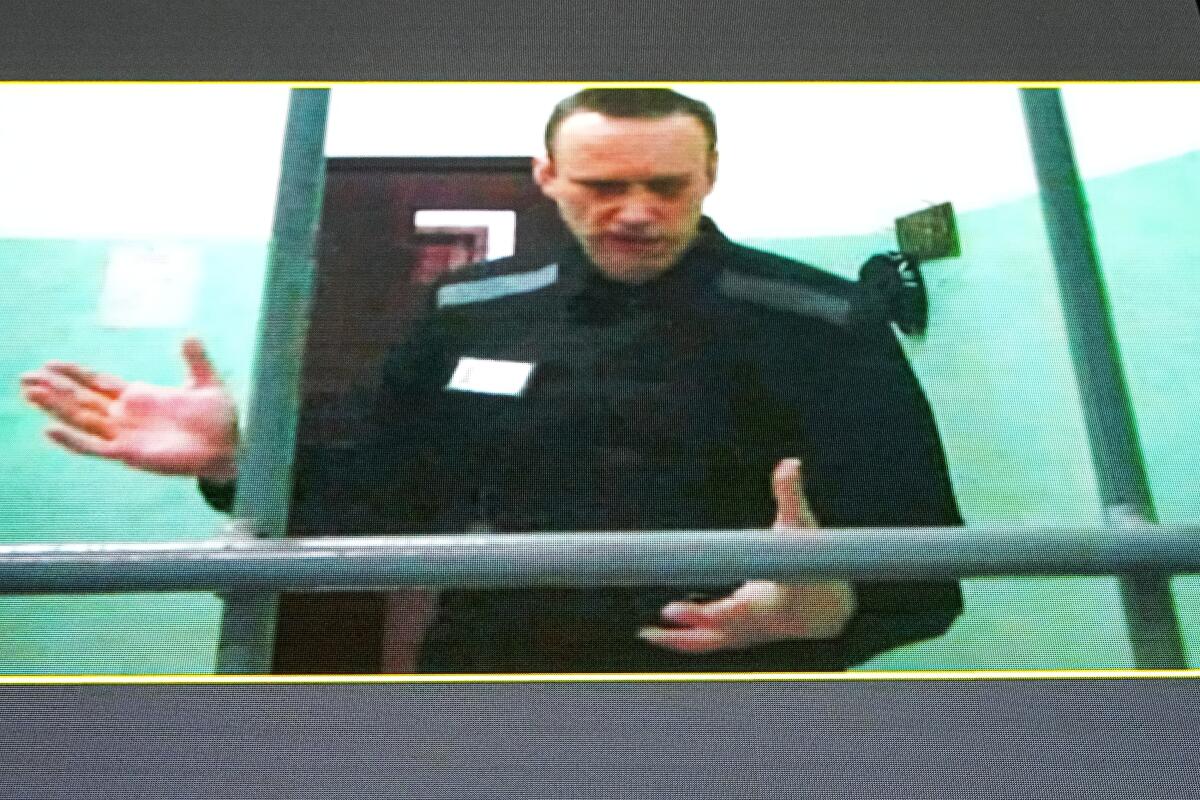 A man in a black uniform gesturing as he speaks from inside a caged-off space indoors.