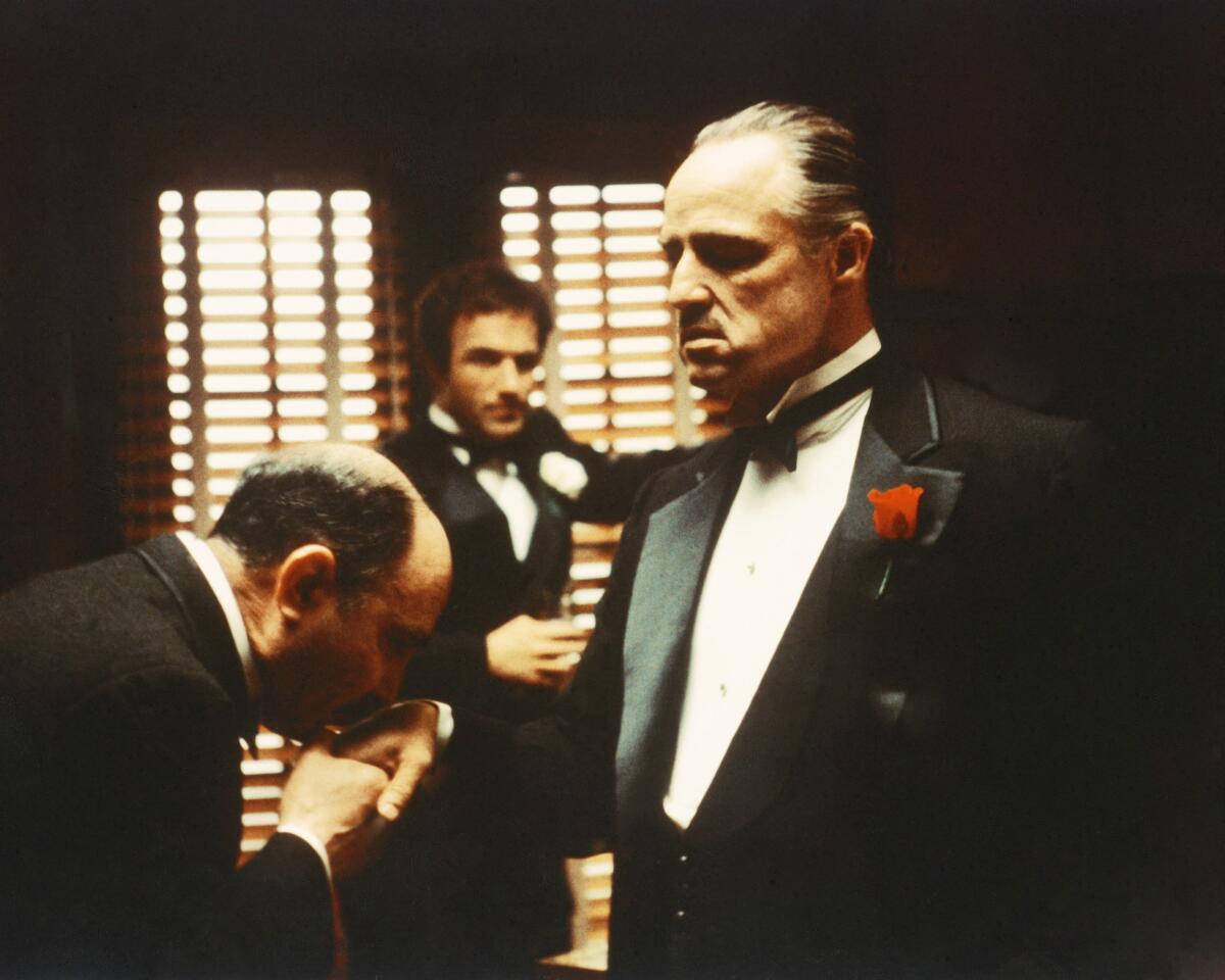 A man kisses the hand of another man as a third man in the background looks on in a scene from "The Godfather."