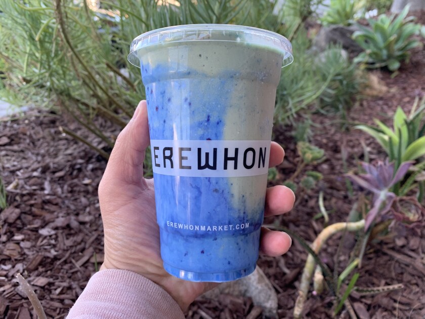 A hand holds a plastic cup with the word Erewhon, which contains a blue and white smoothie.