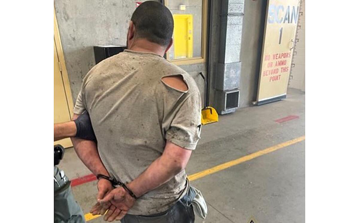 A man, seen from behind, has his hands cuffed behind his back. He wears a T-shirt torn at one shoulder.