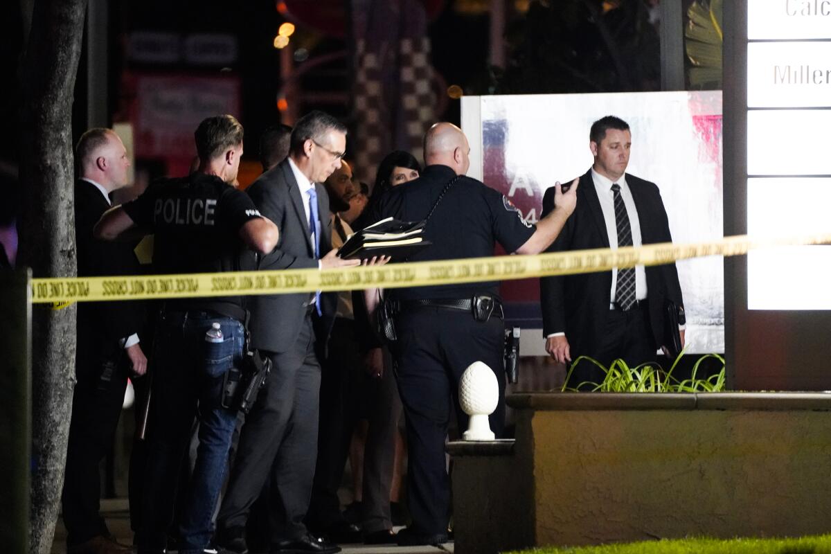 Police officers and men in suits appear behind a yellow police tape.