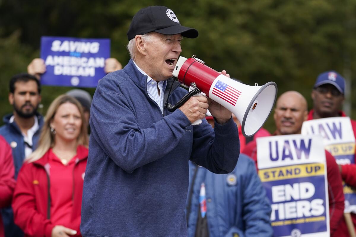 President Biden, in a windbreaker and ballcap, speaks into a bullhorn to people, some holding signs including "UAW stand up"