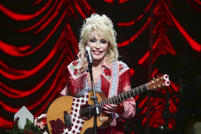 A woman with voluminous blond hair holding a guitar and standing in front of a microphone on a stage