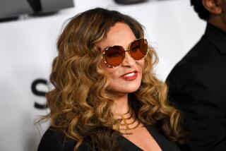 Tina Knowles in a black dress, big sunglasses and a gold necklace smiling and posing against a white background