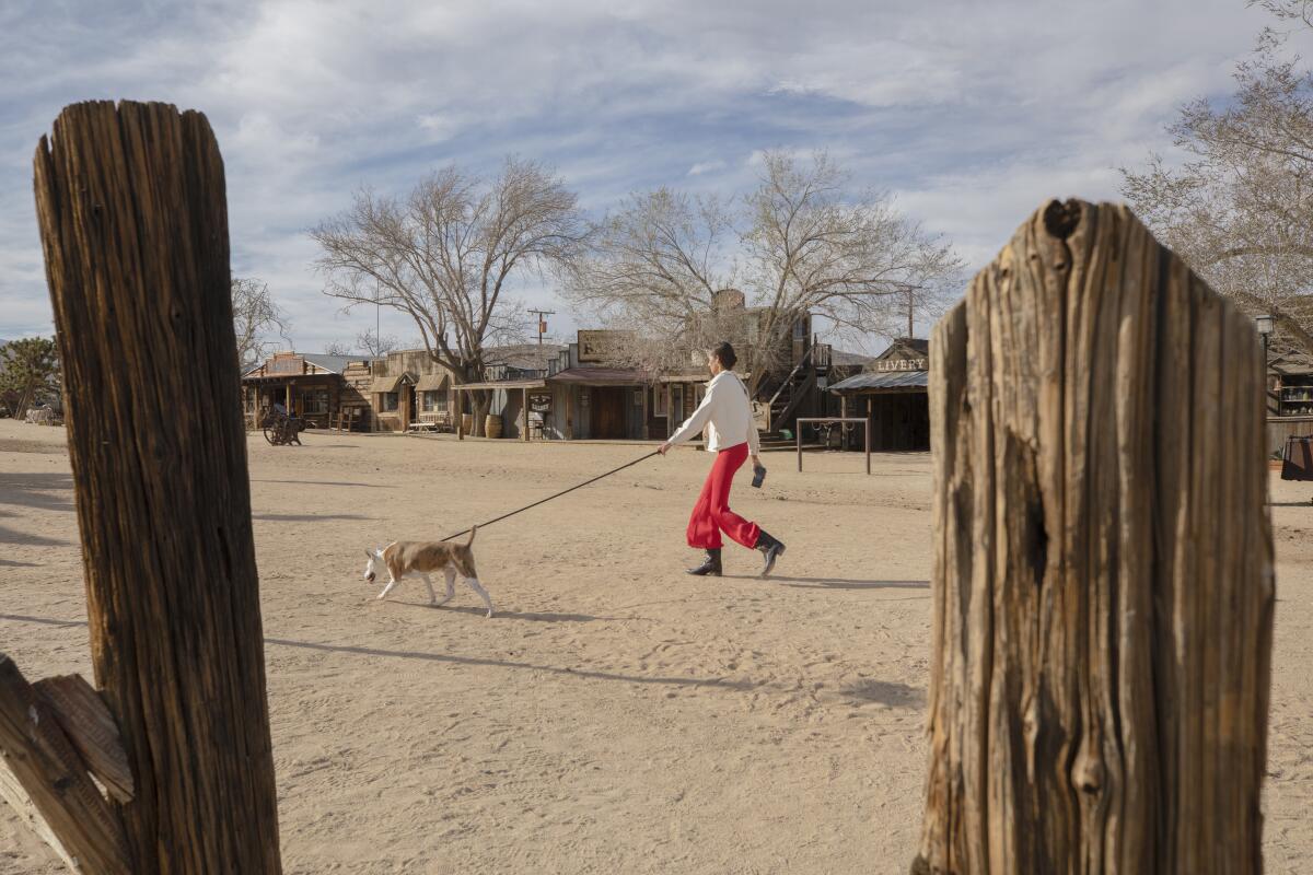 A person walks a dog across a desert expanse in front of old wooden buildings.