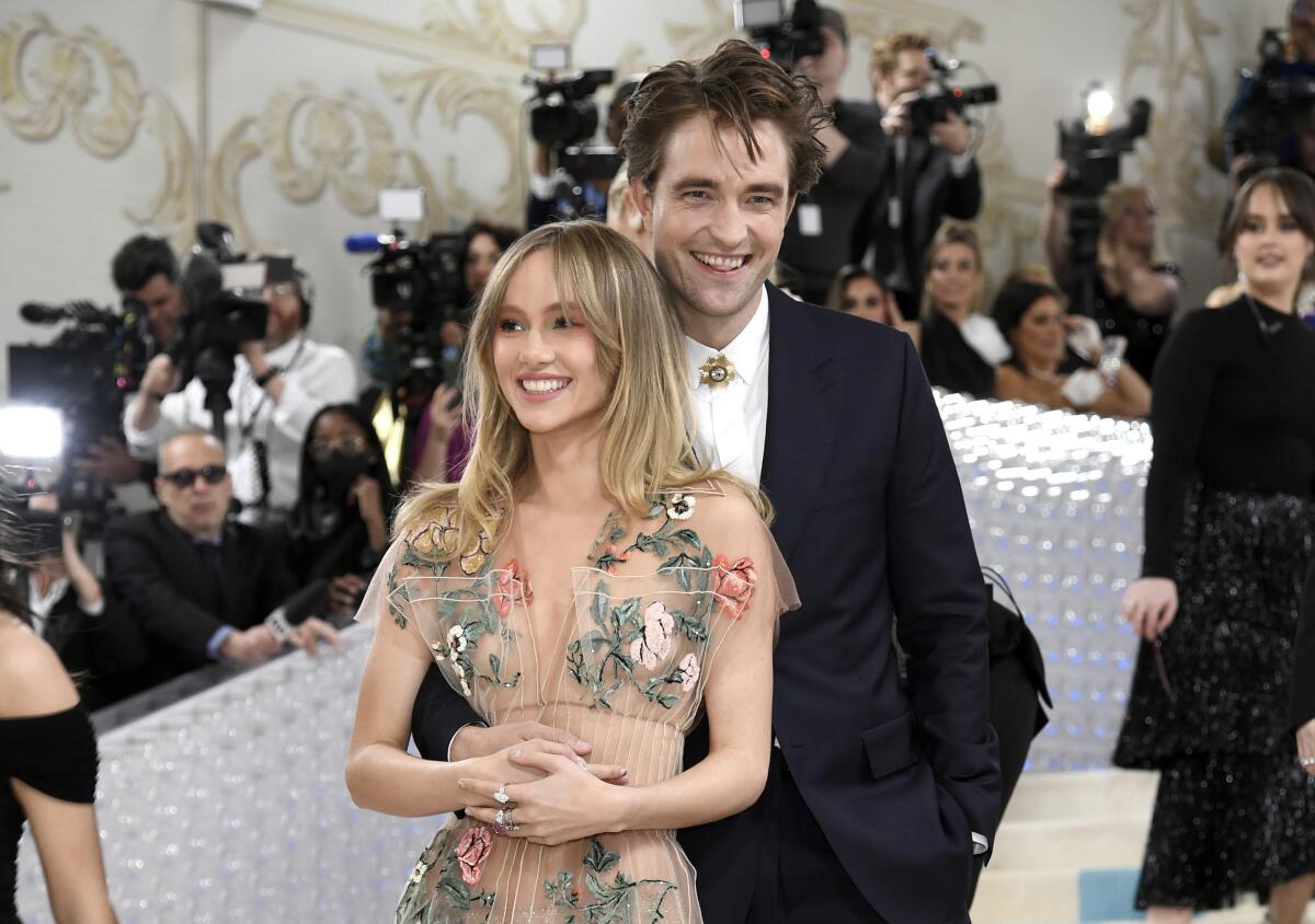 Suki Waterhouse smiles at a gala and clasps her hands  as Robert Pattinson, behind her, puts one arm around her waist