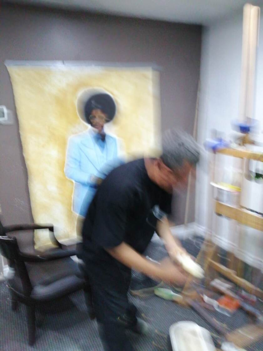 A blurry view of a man with his back to a painting in progress of a Black man in a blue suit against a mustard background