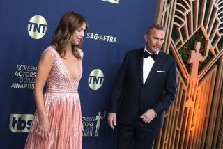 Kevin Costner is wearing a black tuxedo and is standing a few feet from Christine Baumgartner, wearing a peach dress
