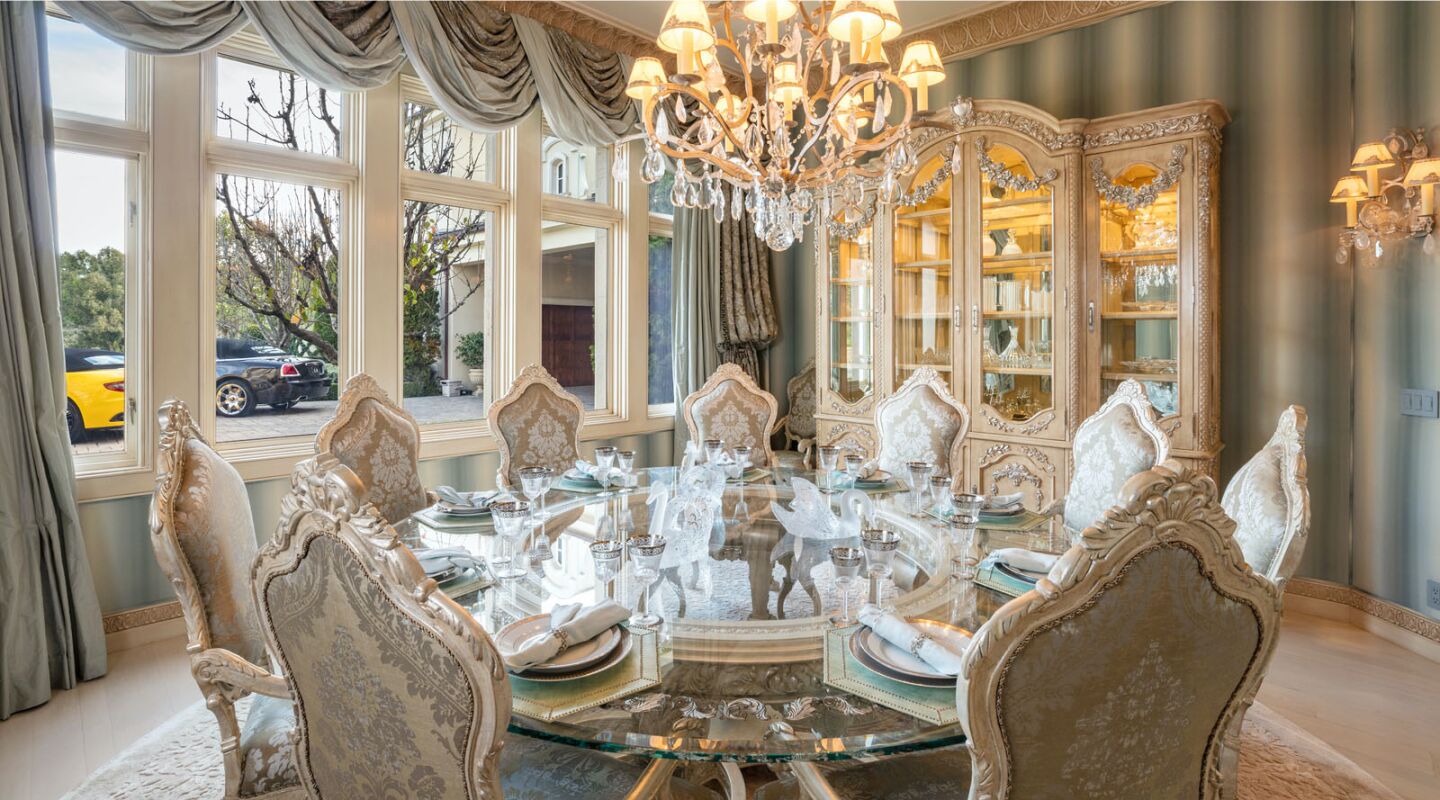 The dining room has elaborate draperies and a chandelier above a large round glass table.