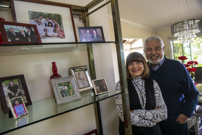 An older couple stands and smiles next to a shelf with framed photos 