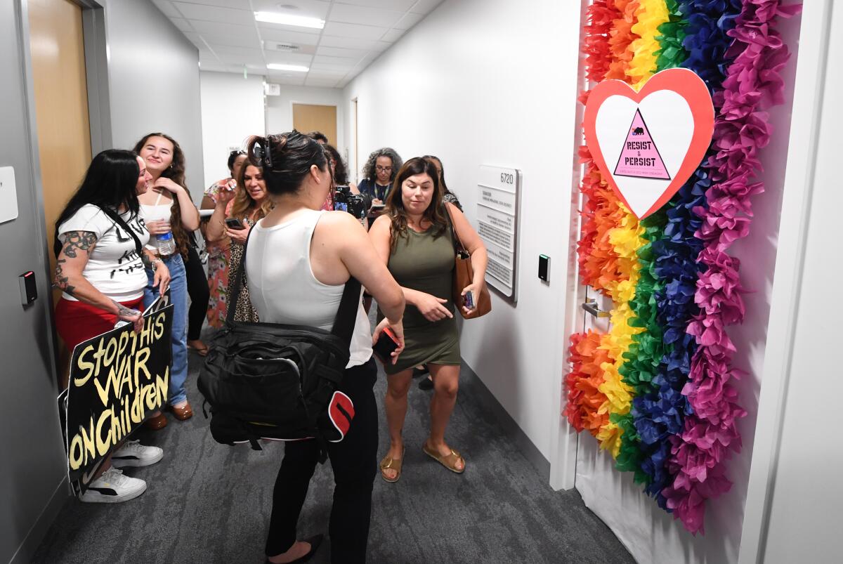 A group standing in a hallway outside an office door covered in a pride flag of fake flowers and a "Resist and persist" sign