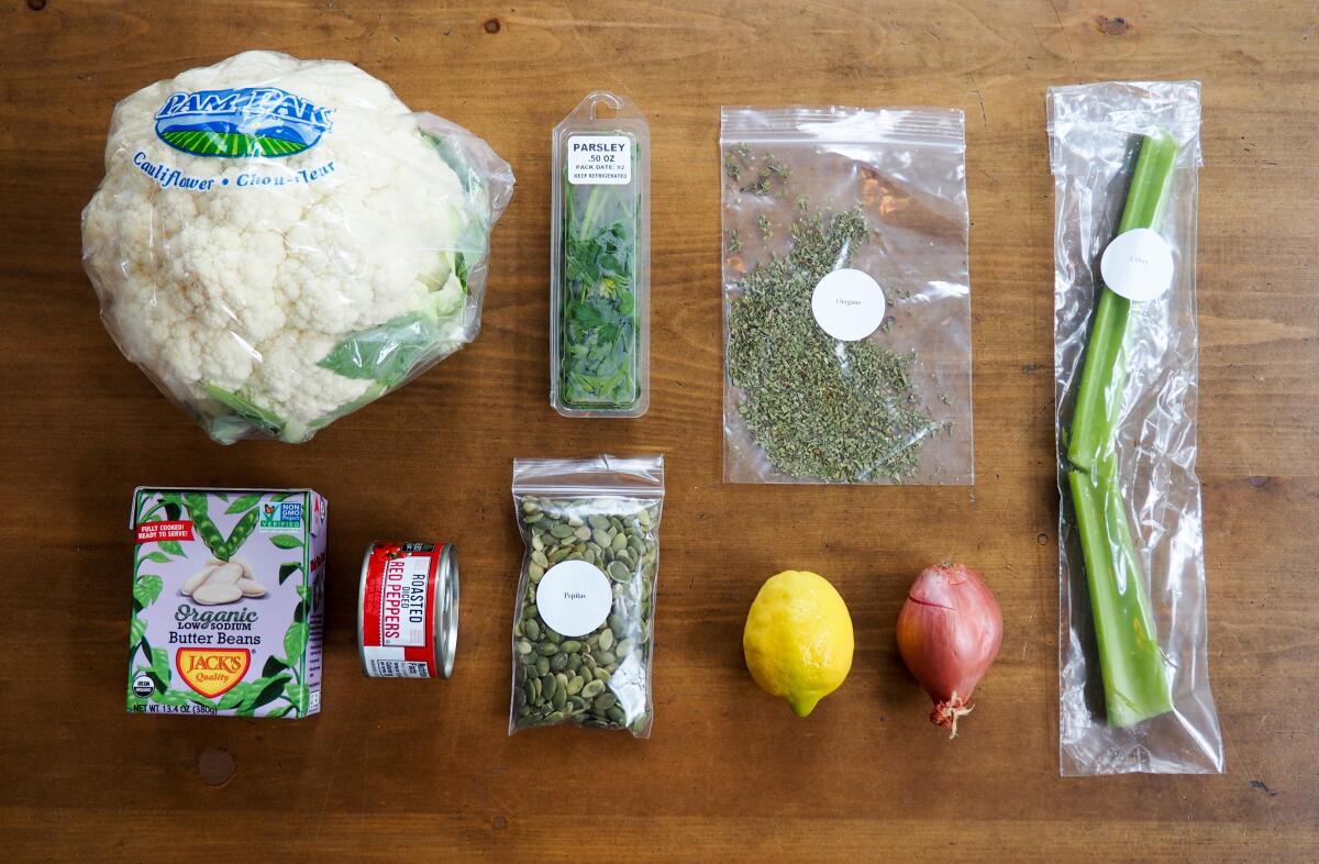 A meal kit box, including cauliflower, beans, lemons, celery and roasted diced red peppers, is laid out on a wooden table.