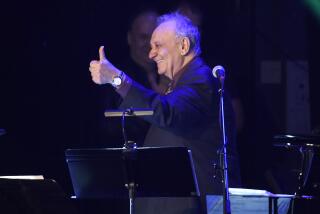 An older, smiling man looks to the side and gives the thumbs-up sign during a musical performance