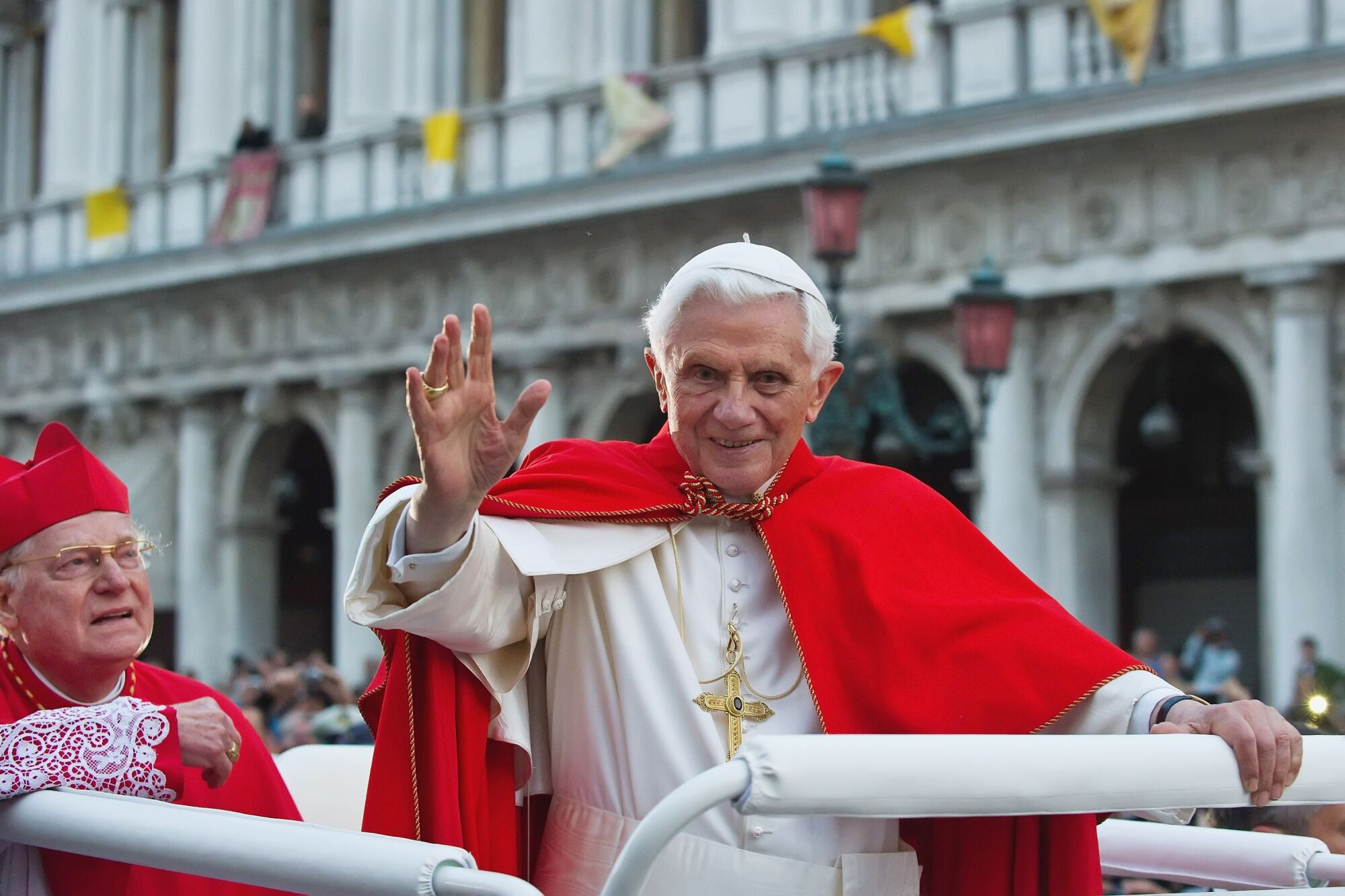 Pope Benedict XVI  waves from a vehicle while another passenger looks on as they drive past a building.