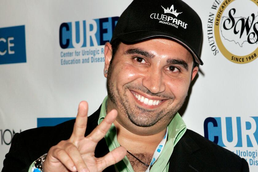 A man, framed waist and up, flashes a hand sign while smiling and posing for the camera.