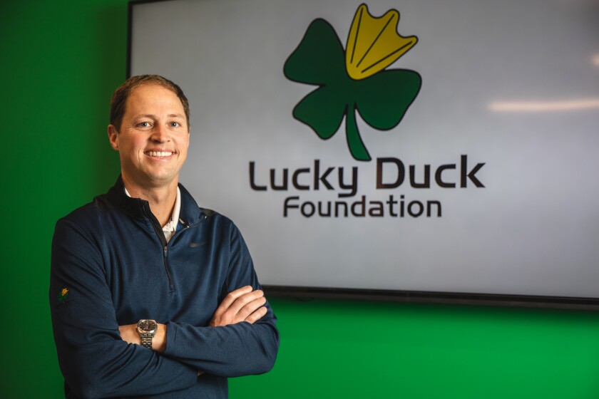 Drew Moser smiling and arms crossed, standing in front of a green wall with Lucky Duck Foundation name and logo 