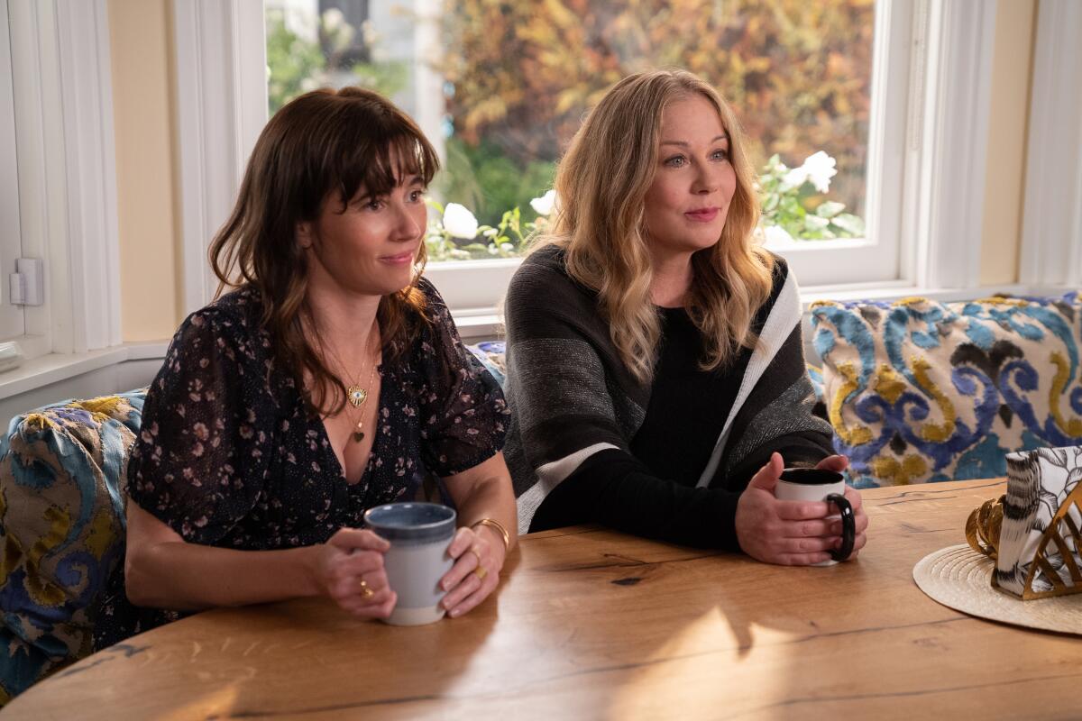 Linda Cardellini and Christina Applegate sit at a table holding coffee cups in a scene from "Dead to Me."