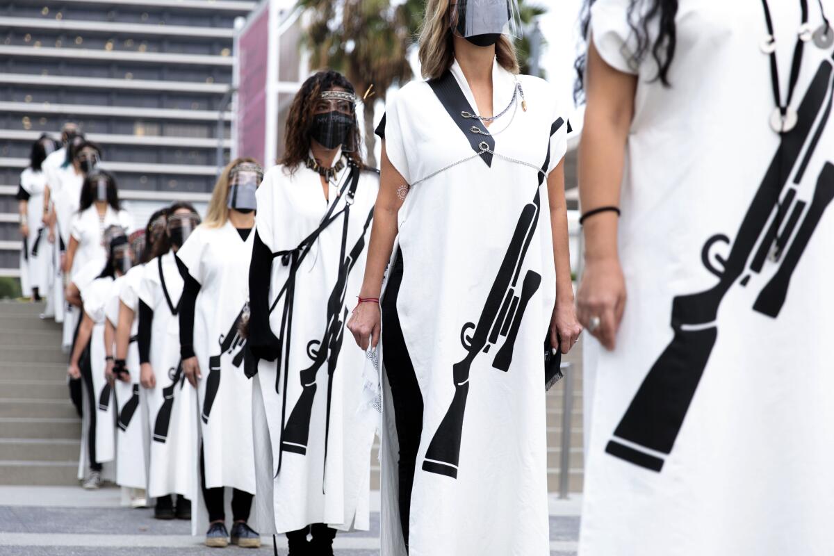 A row of women in white gowns bearing the image of a rifle walk along a downtown Los Angeles street