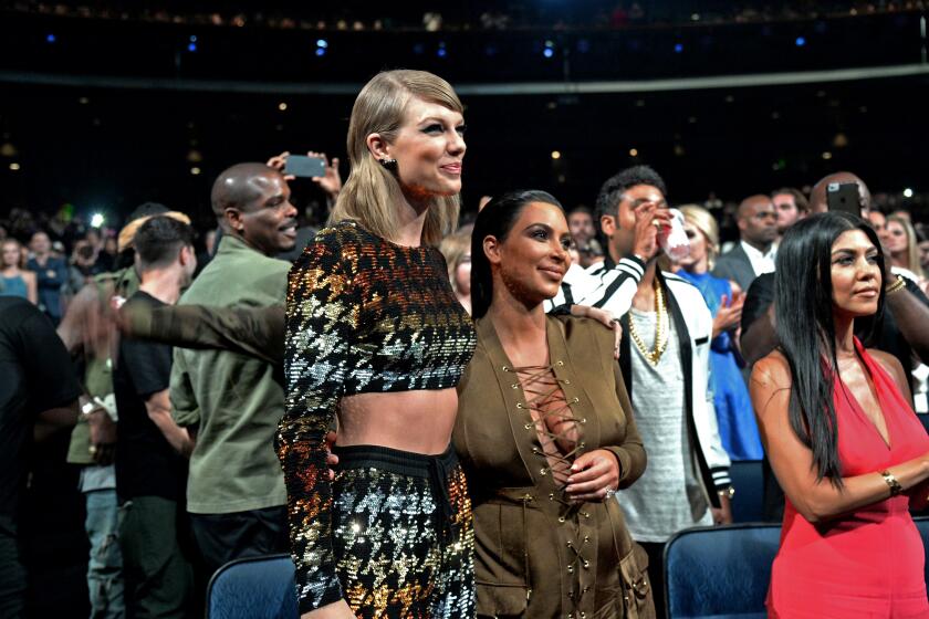 Taylor Swift, in multicolor matching set, and Kim Kardashian, in green dress,  stand arm in arm in crowd