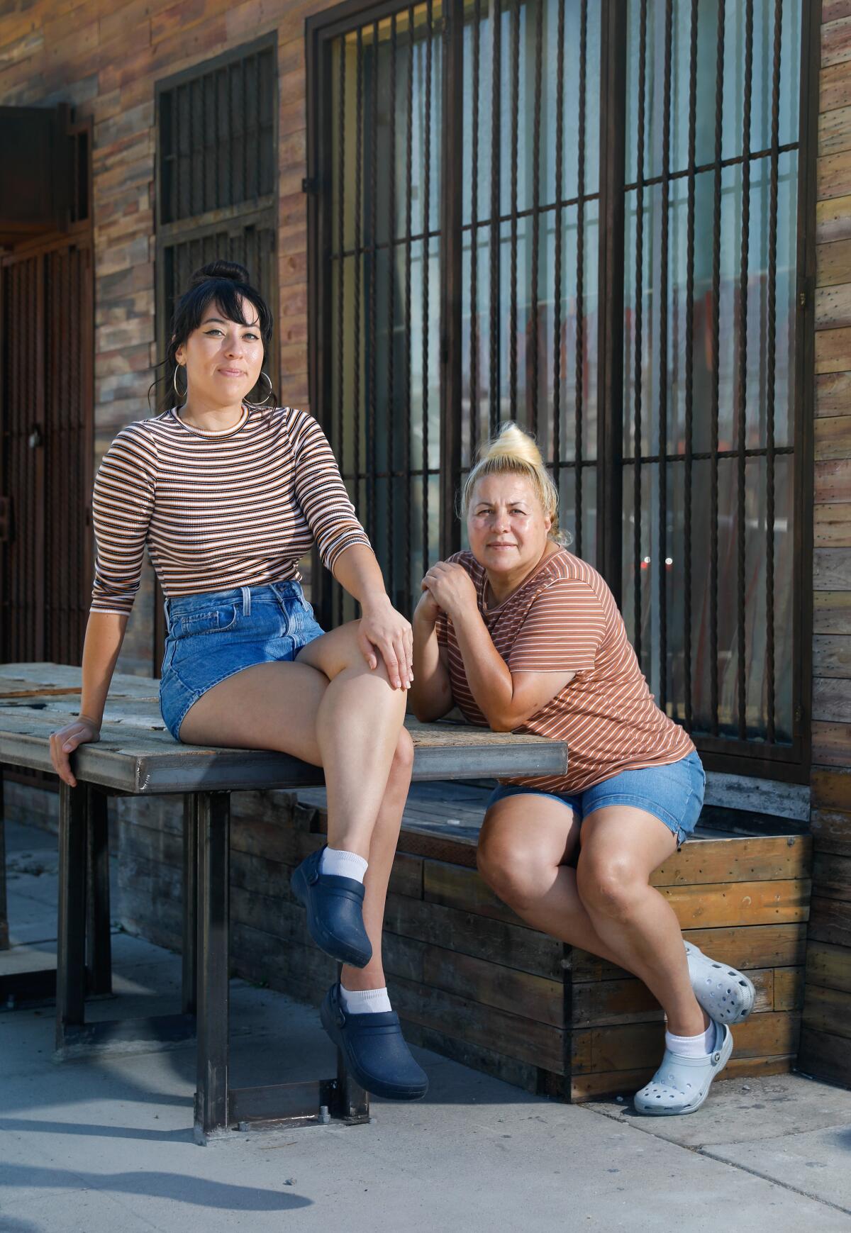 A woman seated on a table and a woman seated on its bench, both wearing Crocs, striped shirts and jean shorts.
